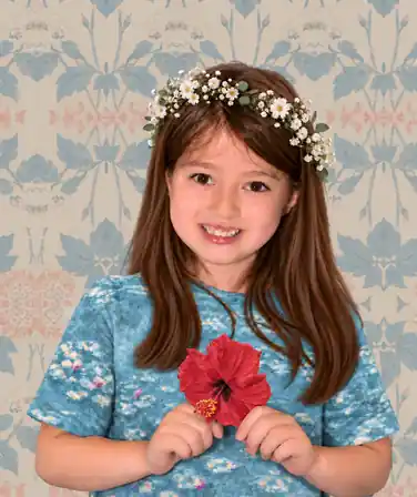 Lillian smiling beneath a white floral crown, with a red hibiscus over her hands and a softly patterned floral background.