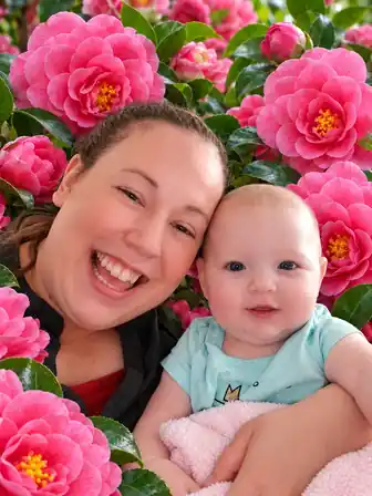 A smiling mother holding her baby, surrounded by pink camellia blossoms and green leaves.