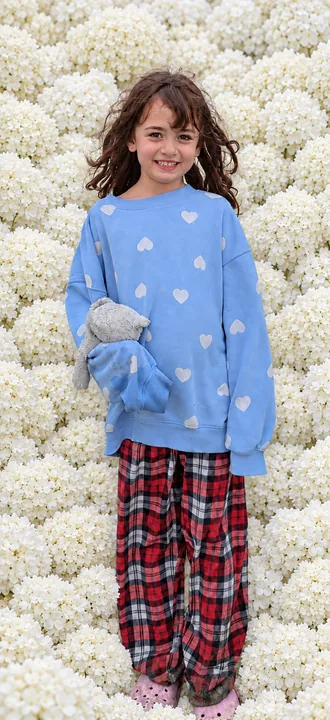 A smiling girl in a blue heart-patterned sweatshirt holding a plush toy, surrounded entirely by white hydrangea blossoms.