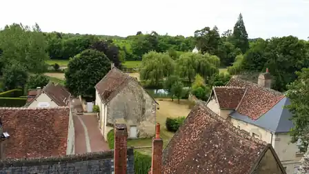 Weathered terracotta and slate village rooftops and chimneys above an empty lane, with a pond and weeping willows beyond.